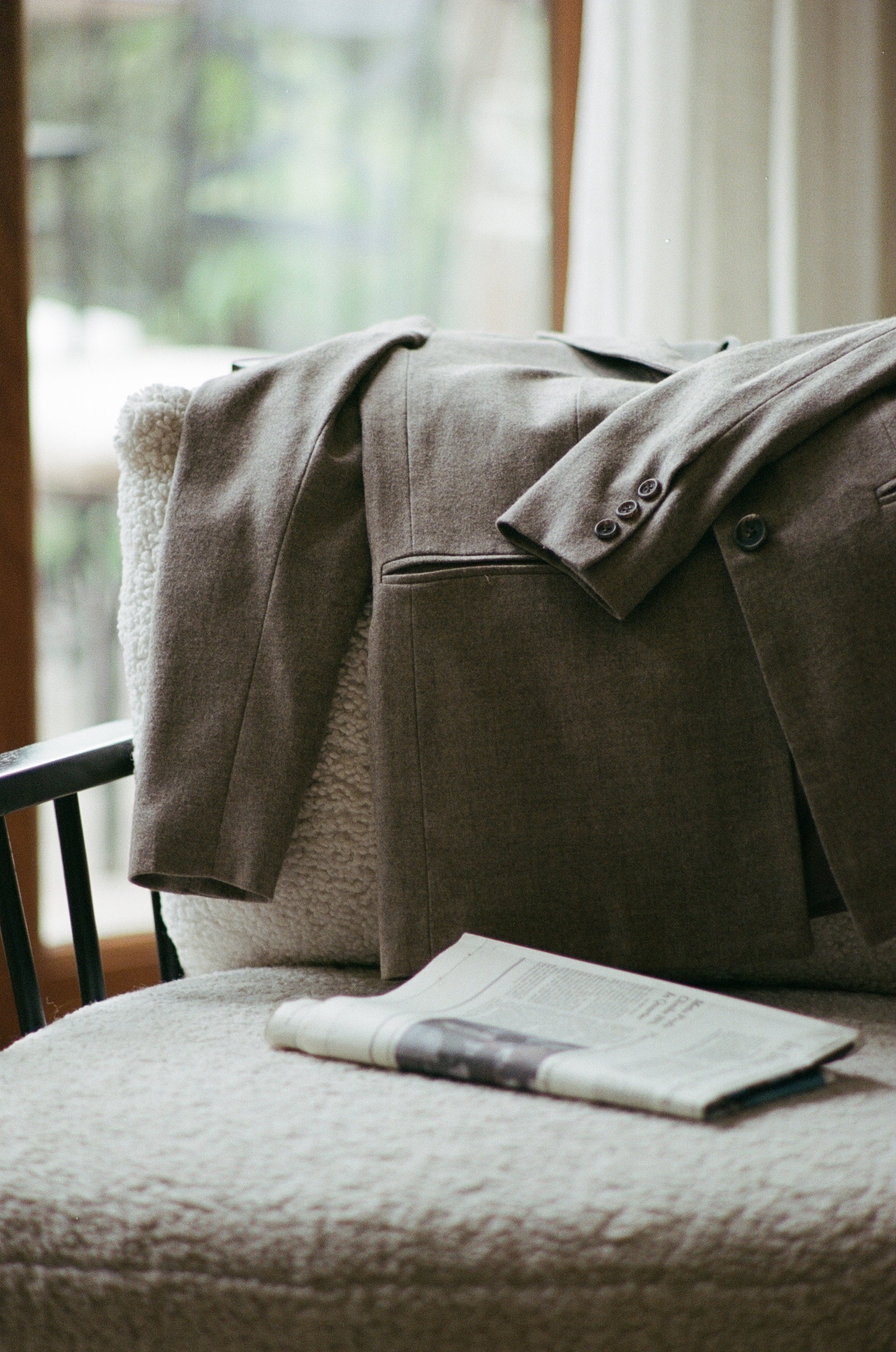 Court Jacket in Coffee hanging over the back of a sherpa lounge chair with a newspaper placed on cushion